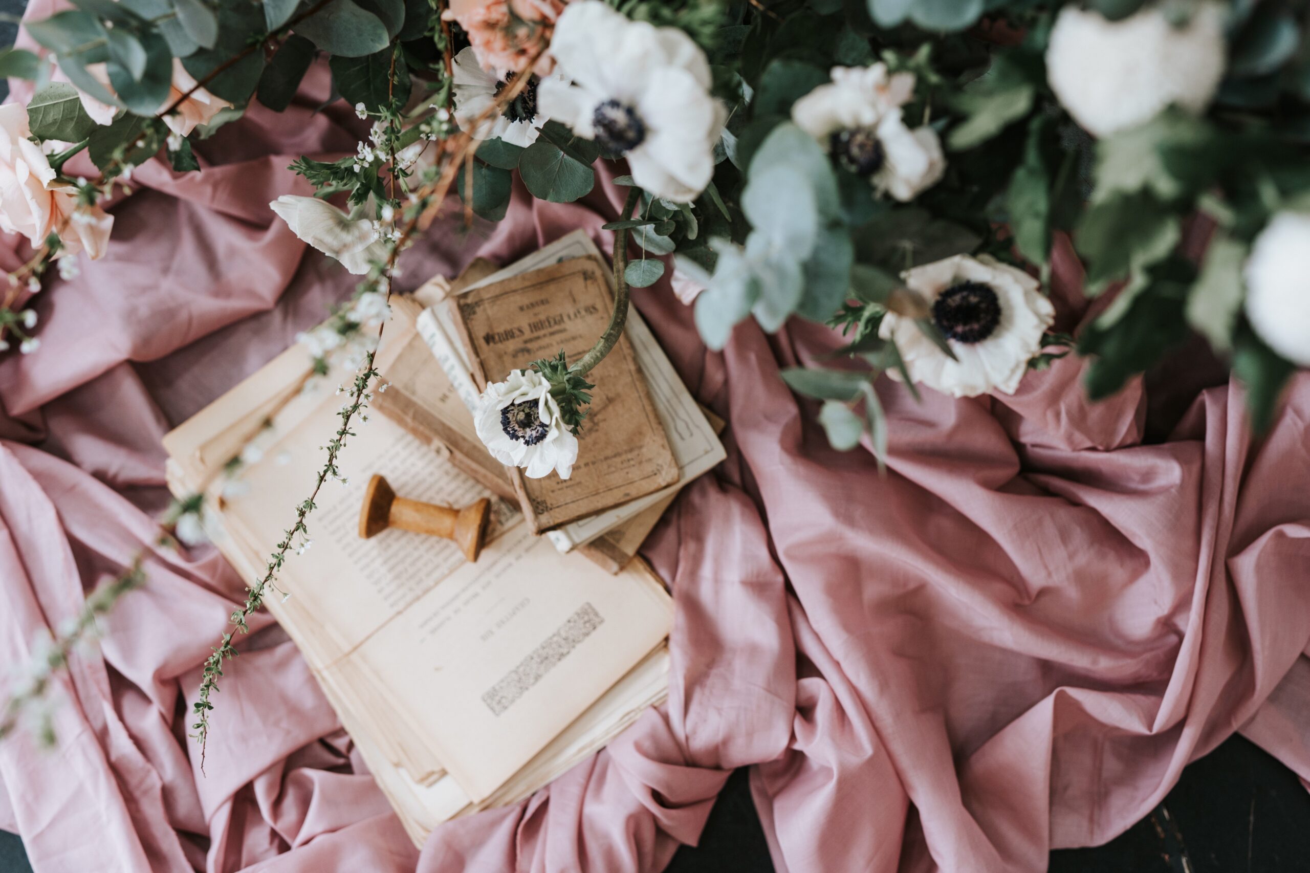 Vintage papers and wooden stamp strewn on a dusty pink cloth with eucalyptus and white anemone flowers in the foreground