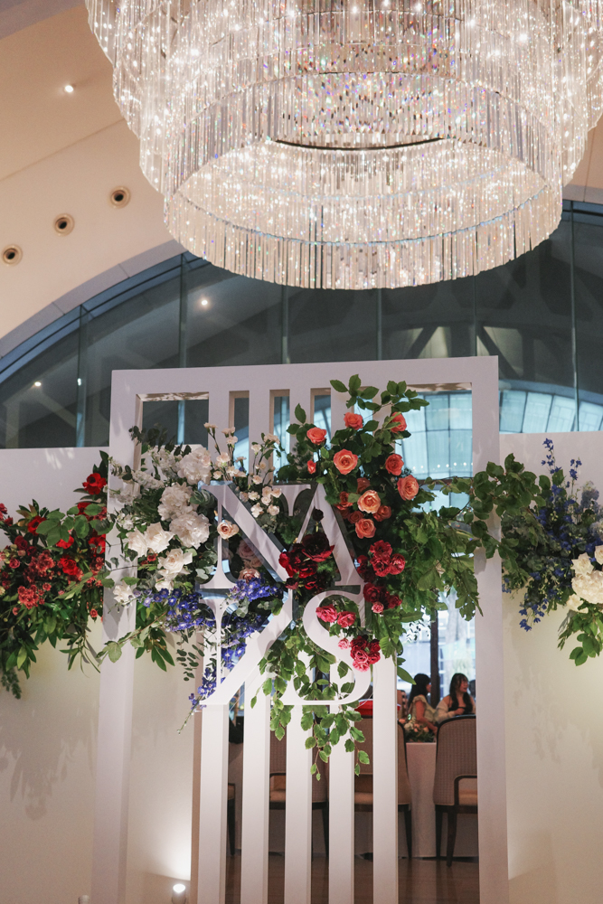 An elaborate overhead floral installation for the wedding entrance with the couple's initials, featuring white, peach, and red flowers and foliage, consisting of Roses, Hydrangeas, Delphiniums, Snapdragons, Peonies, Lisianthus and Dianthus suspended beneath a modern chandelier at The Clifford Pier at The Fullerton Bay Hotel.