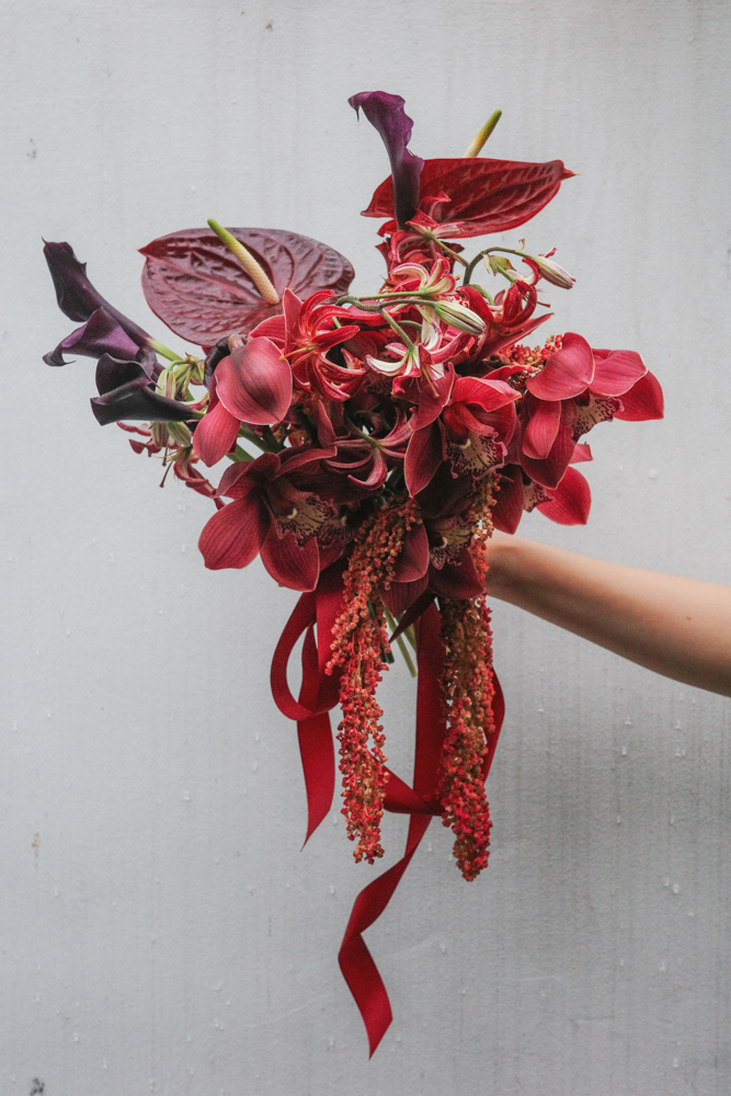 A hand gently holds a vibrant and textured bouquet featuring deep crimson orchids, velvety anthuriums, burgundy calla lilies, and striking gloriosa lilies, cascading with amaranthus and wrapped in trailing red silk for a wedding bouquet with trailing ribbons in matching hues at The Clifford Pier at The Fullerton Bay Hotel.