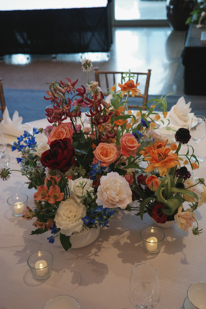 A vibrant and textured centerpiece arrangement with orange, blue, white, and burgundy flowers, consisting of Delphiniums, Tiger Lilies, Roses, Carnations, Forget Me Nots and Tweedia in a clear vase, placed on a light-colored table with candles at The Clifford Pier at The Fullerton Bay Hotel.