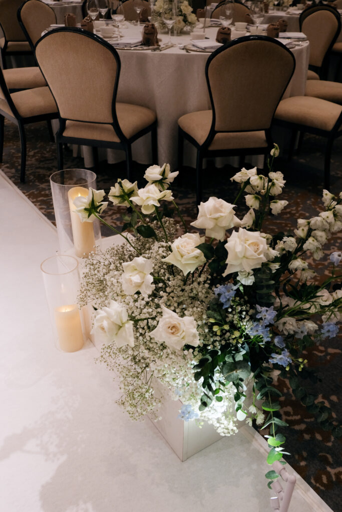 A floor-level view of elegant white floral arrangements with touches of greenery, consisting of Roses, Baby Breaths, Delphiniums and Orchids, placed near round tables with chairs in a wedding celebration at Raffles Hotel Singapore.