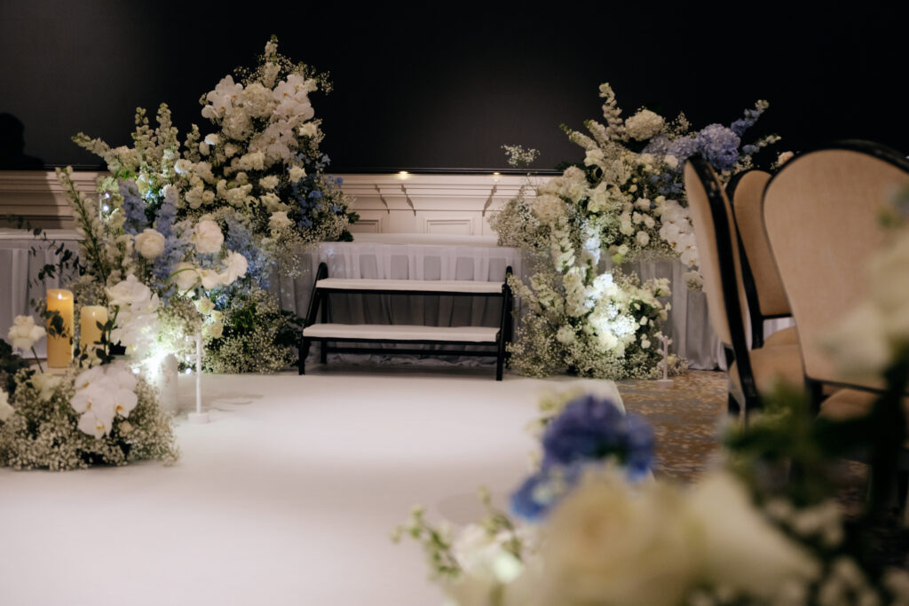 A beautifully decorated stage with white floral on the sides of the stairs, consisting of Roses, Baby Breaths, Delphiniums, Hydrangeas and Orchids, with soft lighting, and neutral-toned seating at Raffles Hotel Singapore.