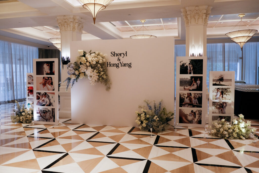 A photo area backdrop from the wedding entrance with a patterned floor, decorated with white floral arrangements along a pathway and a backdrop featuring signage and photos, consisting of Roses, Baby Breaths, Delphiniums, Hydrangeas and Orchids at Raffles Hotel Singapore.