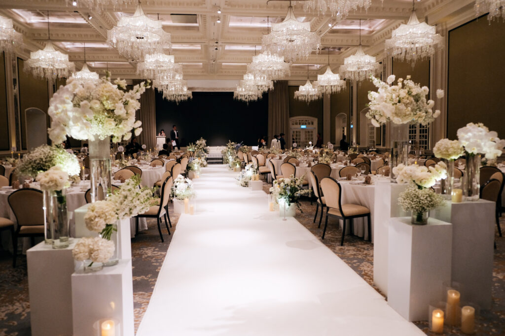 A long, elegant wedding aisle with a white runner, flanked by tall white floral arrangements on pedestals, consisting of hydrangeas, roses, baby breaths and orchids leading towards a decorated area with soft, overhead lighting at Raffles Hotel Singapore.