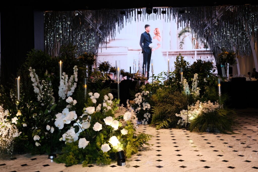 Elegant wedding stage decoration in Singapore featuring lush white floral arrangements, consisting of delphiniums, roses, and lilies with candles, and a softly lit backdrop where the couple photos together on the screen at JW Marriott Hotel Singapore South Beach.