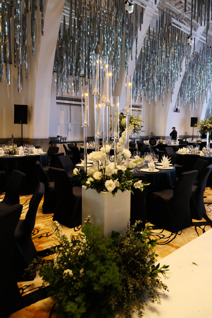 A wedding reception table setting with elegant black chairs and tables, white tablecloths, and a tall centerpiece featuring white flowers and greenery, consisting of roses and delphiniums, illuminated by warm lighting in a sophisticated venue at JW Marriott Hotel Singapore South Beach.