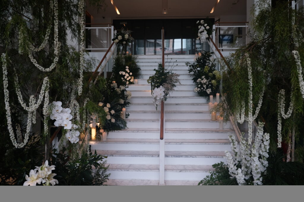 A grand staircase adorned with lush white and green floral consisting of jasmine floral draping, phalaenopsis orchids, lilies, delphiniums and roses, leading upwards in a formal wedding venue at JW Marriott Hotel Singapore South Beach.