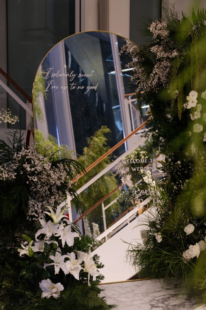 Close-up of a uniquely decorated wedding entrance through mirror reflections, featuring a round mirror with calligraphy or signage, surrounded by asymmetrical arrangements of white and natural-toned flowers and foliage at JW Marriott Hotel Singapore South Beach.