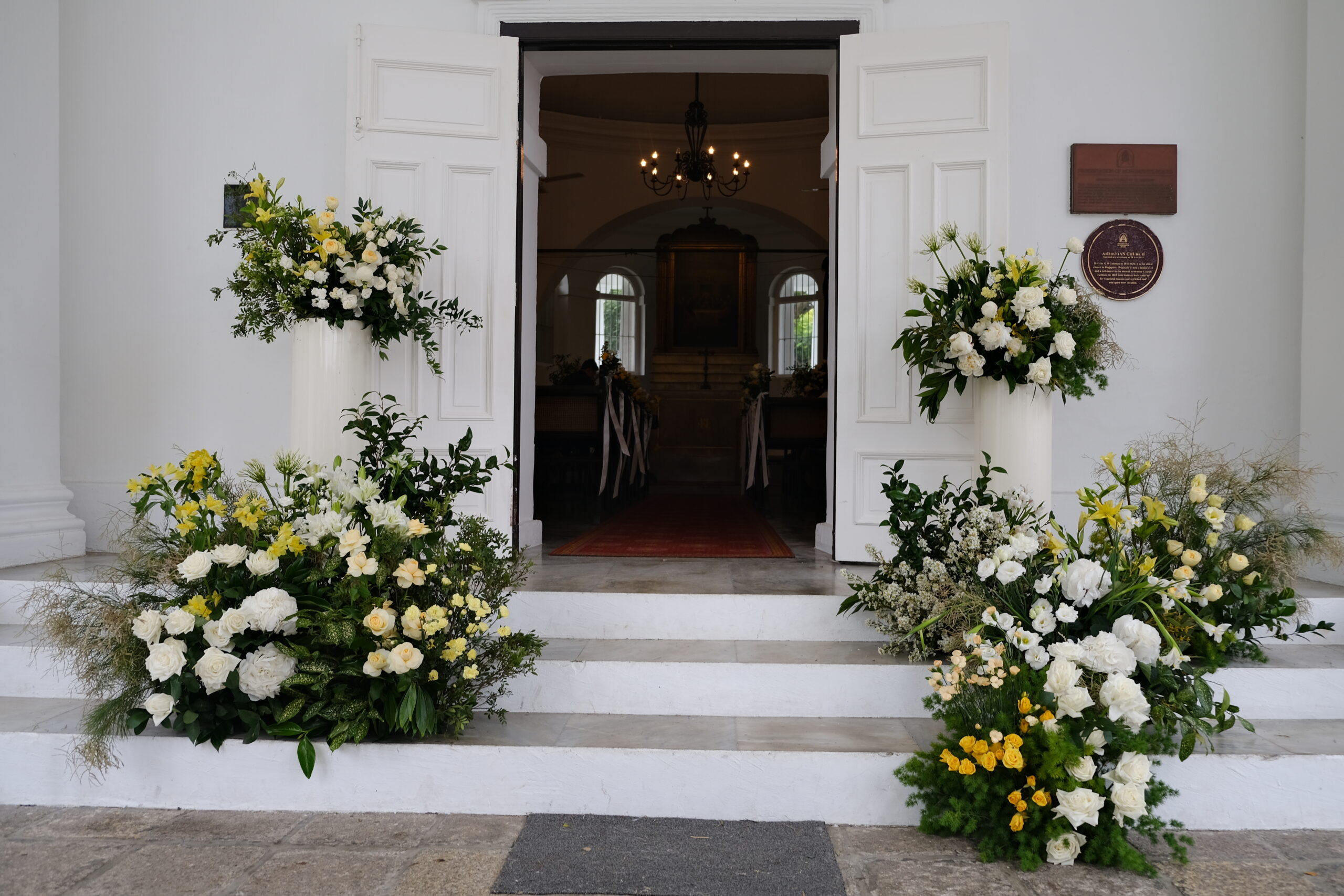The entrance of a church adorned with white and yellow flowers consisting of Roses, Lilies, and Lisianthus displayed on pedestals and along the steps, is prepared for a wedding celebration at the Armenian Church in Singapore.