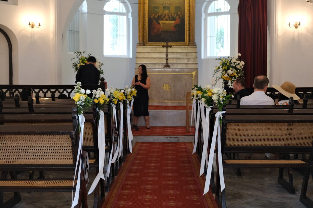 The interior aisle of a church decorated for a wedding with a white and yellow roses arrangement with greenery attached to the pews and white ribbons marking the walkway at the Armenian Church in Singapore.