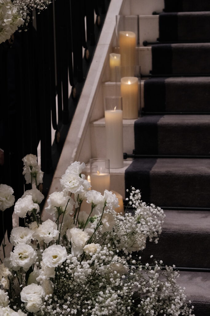 A dimly lit staircase adorned with lush arrangements of white flowers, including lisianthus and baby's breath, and illuminated by scattered candles on the steps for a luxury wedding at Caroline Mansion Singapore.