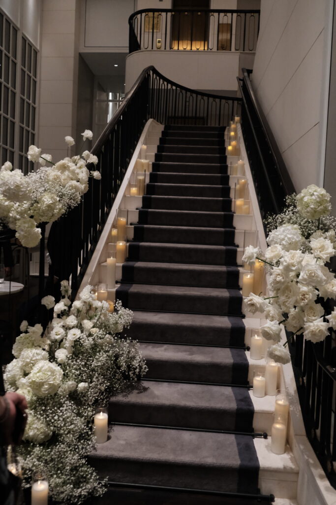 A wide shot of a staircase decorated with abundant white floral arrangements, consisting of fresh white roses, hydrangeas, lisianthus, carnations, and baby’s breath. glowing candles along the steps, creating a romantic and inviting atmosphere in a dimly lit setting for a luxury wedding at Caroline Mansion Singapore.