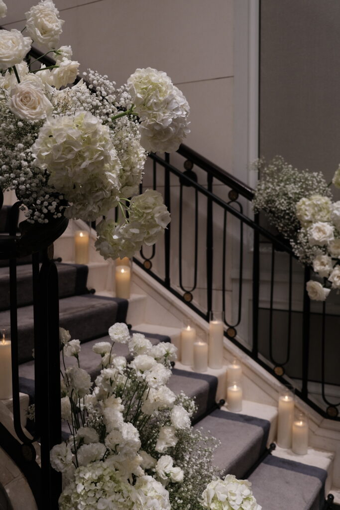 A staircase elegantly decorated for a wedding or event with cascading white flowers, including fresh white roses, hydrangeas, lisianthus, carnations, and baby’s breath, and warm candlelight illuminating the steps and railings for a luxury wedding at Caroline Mansion Singapore.