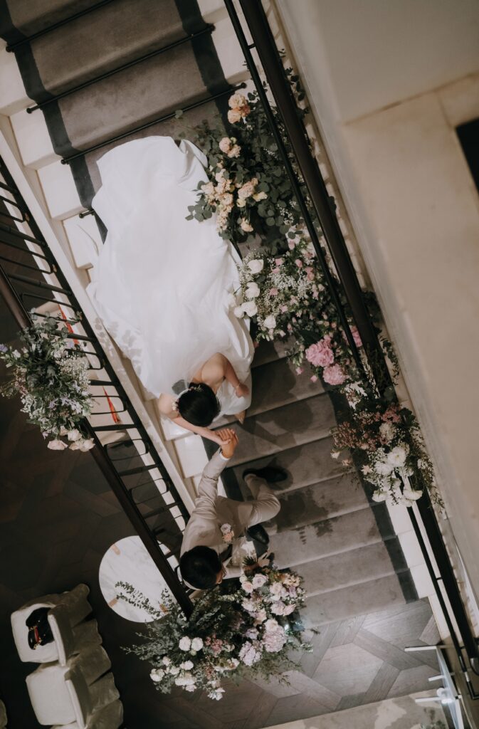 An overhead shot of a newly married couple descending a staircase adorned with elegant floral garlands featuring pastel blooms in white, pink, and purple, accented by lush greenery.