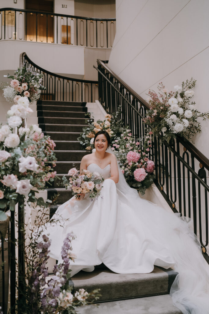A bride in a white off-the-shoulder wedding dress poses gracefully on a staircase embellished with abundant floral decorations in pastel shades of white, pink, and purple, complemented by greenery.
