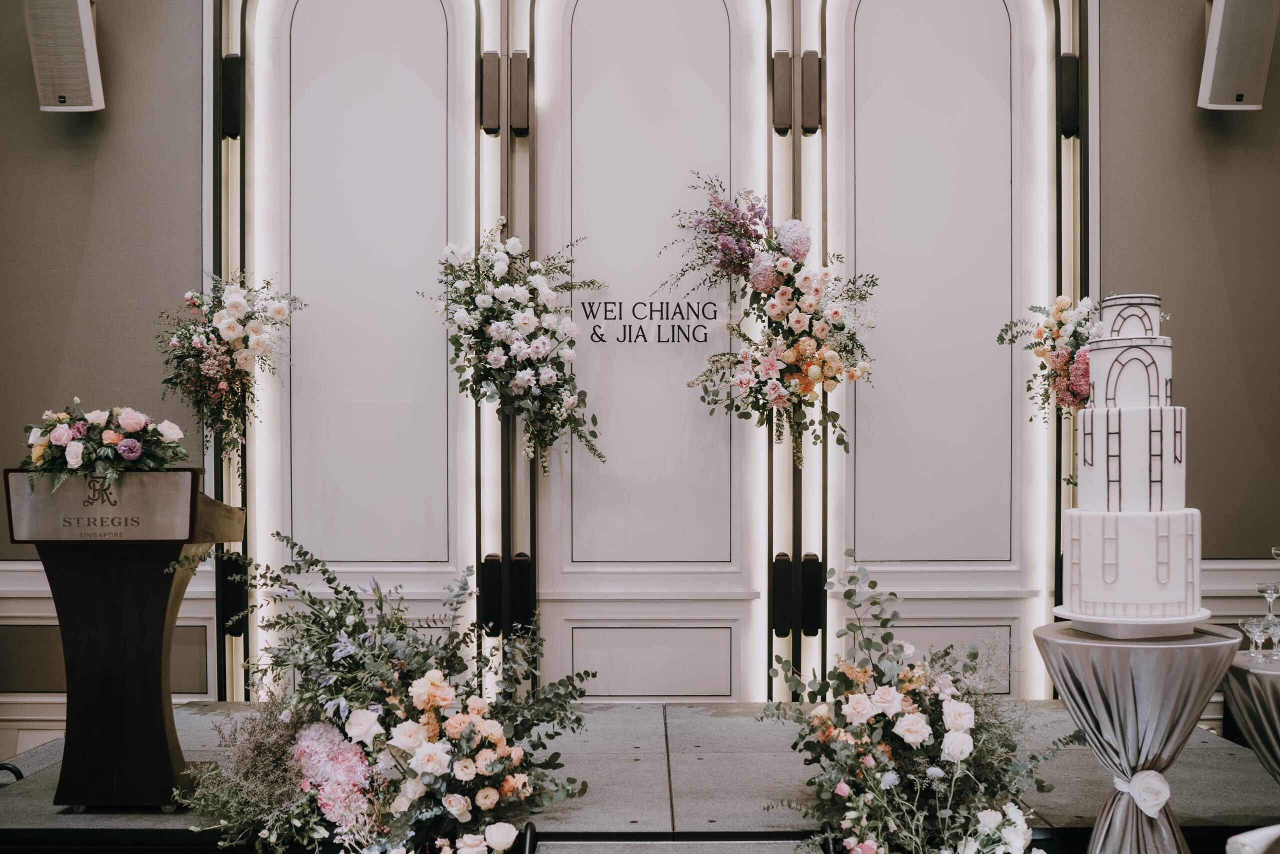 Landscape view of a wedding ceremony or reception backdrop with ornate white panels accented by asymmetrical floral arrangements in shades of white, pink, and purple.