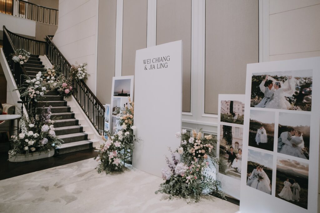 Wedding reception décor featuring a staircase lined with floral arrangements in white, pink, purple, and lush greenery, leading to a display of framed photos and a welcome sign in an elegant font.