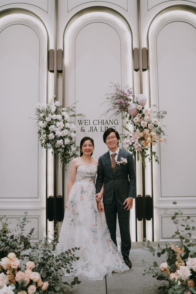 A bride in an elegant white wedding dress and a groom in a dark suit stand smiling in front of a decorated wall featuring a floral backdrop of white, pink, and purple blooms with greenery.