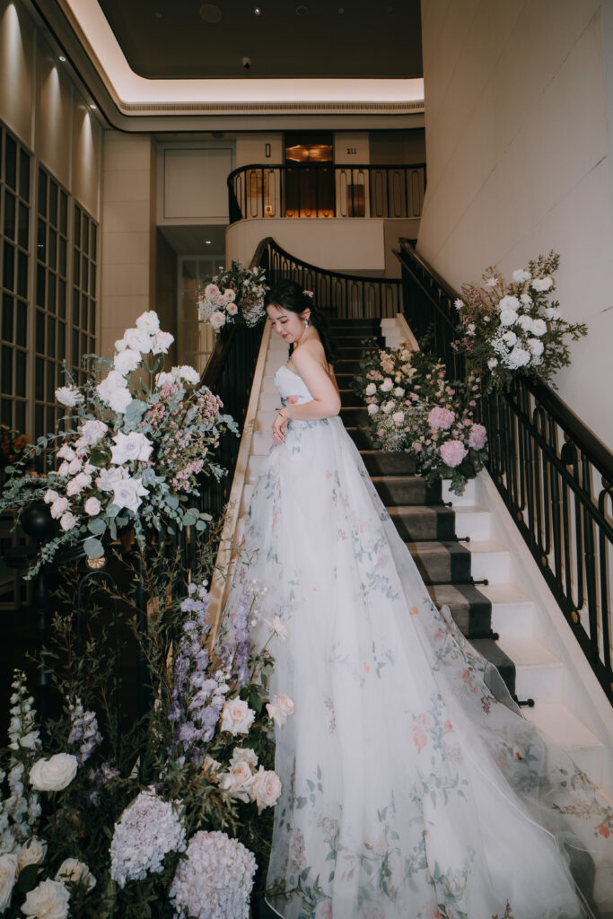A bride in a white lace wedding gown descends a grand staircase adorned with lush floral arrangements in pastel tones of white, pink, and purple, surrounded by foliage.