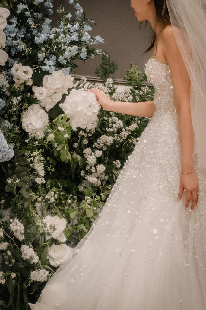 A close-up view of a bride in a white wedding dress, partially visible, standing near lush white floral decorations with delicate greenery consisting of Hydrangeas, Roses, Baby Breaths and Orchids at Raffles Hotel Singapore.