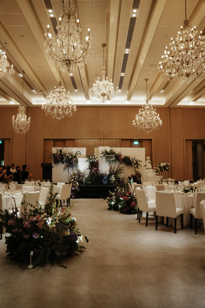 Full view of reception setup with purple theme, garden-style floral designs in white space and chandelier above. 