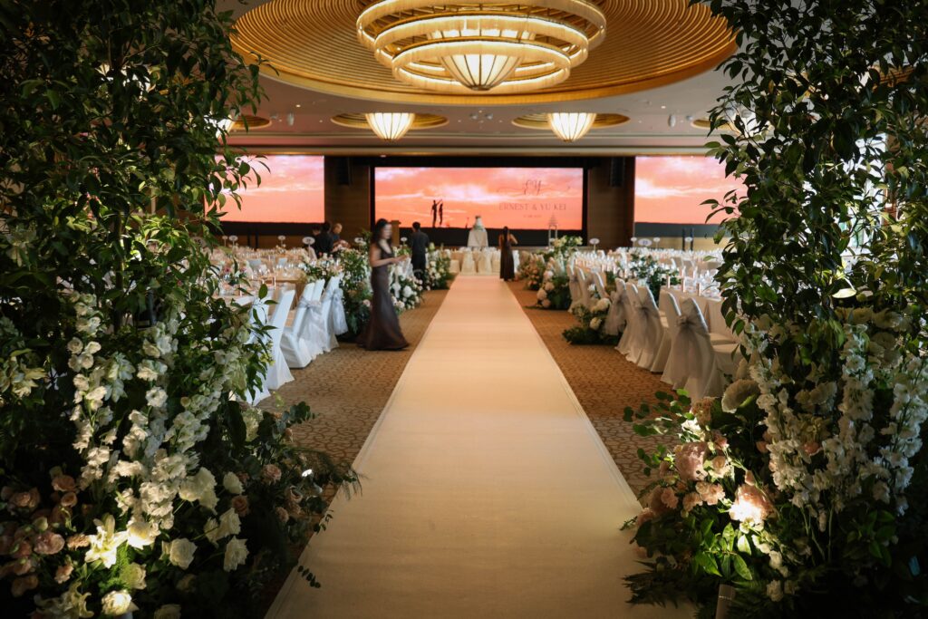 Ceremony entrance framed with tall greenery trees, white hydrangeas and blush roses forming a garden-inspired aisle within the grand ballroom at The Ritz-Carlton Singapore