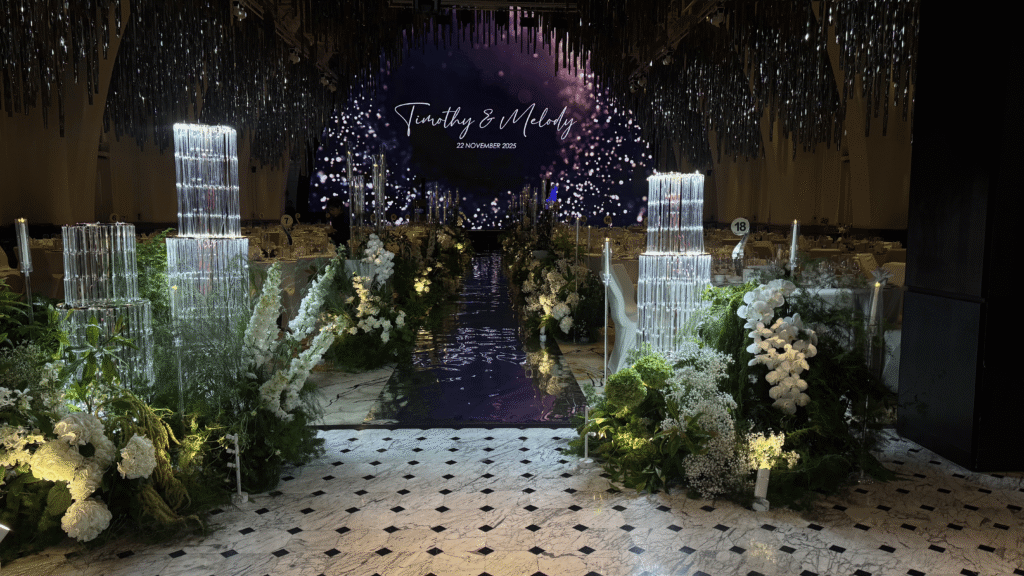 White and green wedding aisle with low lush floral arrangements, candles, and reflective walkway creating a warm glowing entrance at JW Marriott Hotel Singapore South Beach