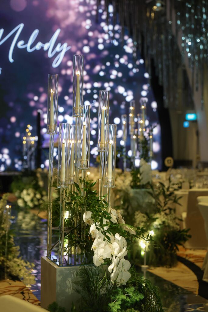 Tall glass cylinder candles surrounded by white flowers and greenery forming vertical accents along the wedding aisle at JW Marriott Hotel Singapore South Beach