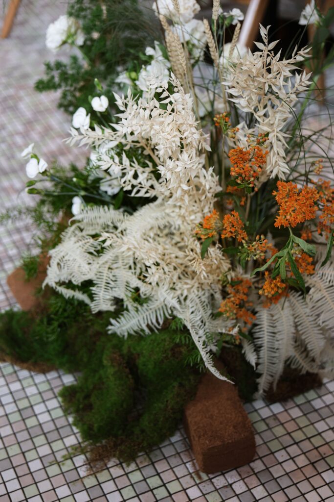 White orchids, soft grasses, textured blooms, and warm earthy florals arranged as a grounded floor installation with wood accents at Min Jiang at Dempsey, Singapore