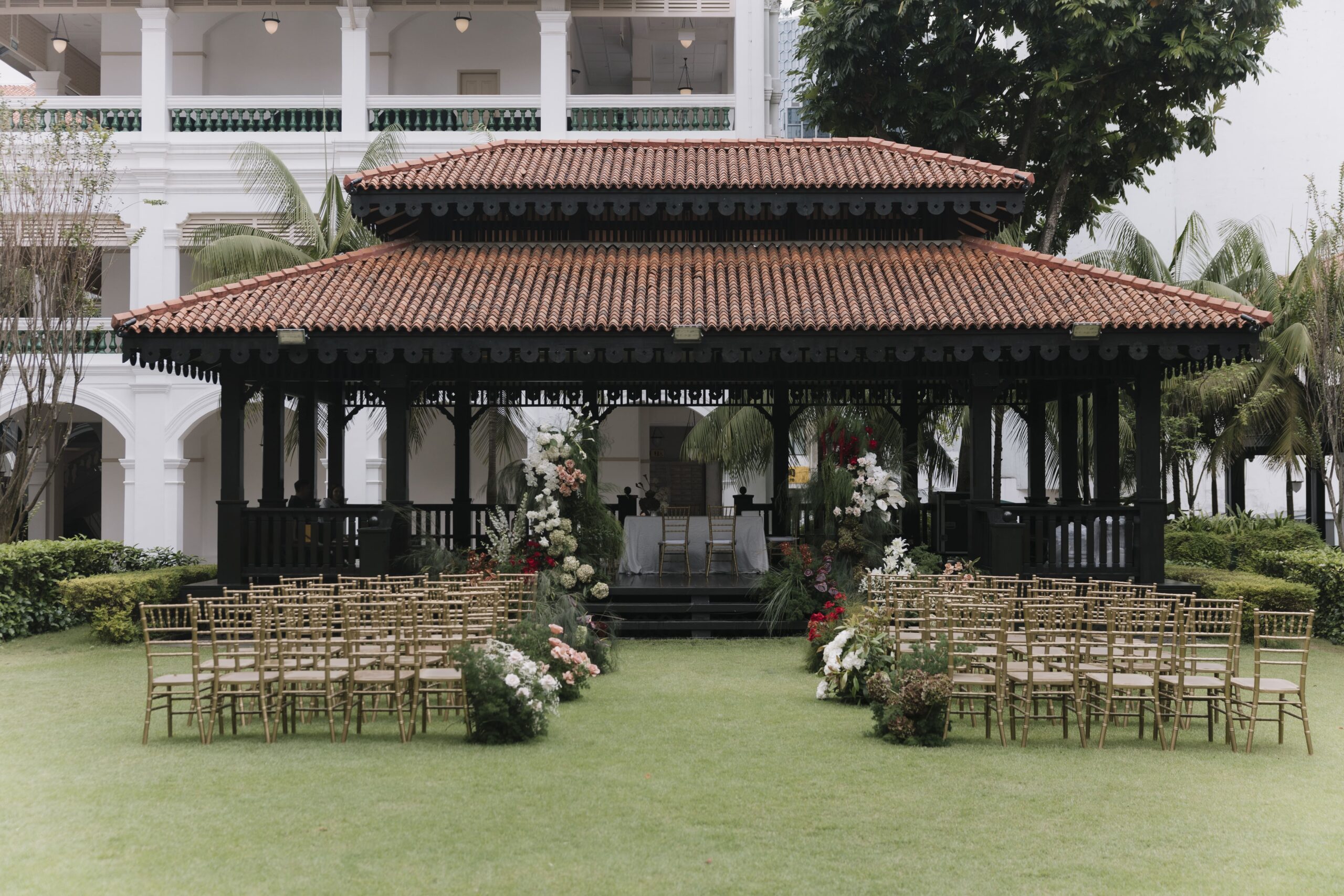 Wide view of colonial pavilion ceremony styling with aisle florals and modern classic lawn wedding design framed by elegant architecture at Raffles Hotel Singapore