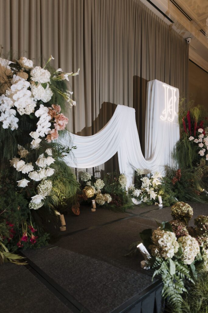 Romantic floral stage-side arrangement with white blooms, blush roses, burgundy accents, and flowing drapery detail for a modern classic ballroom wedding at Raffles Hotel Singapore