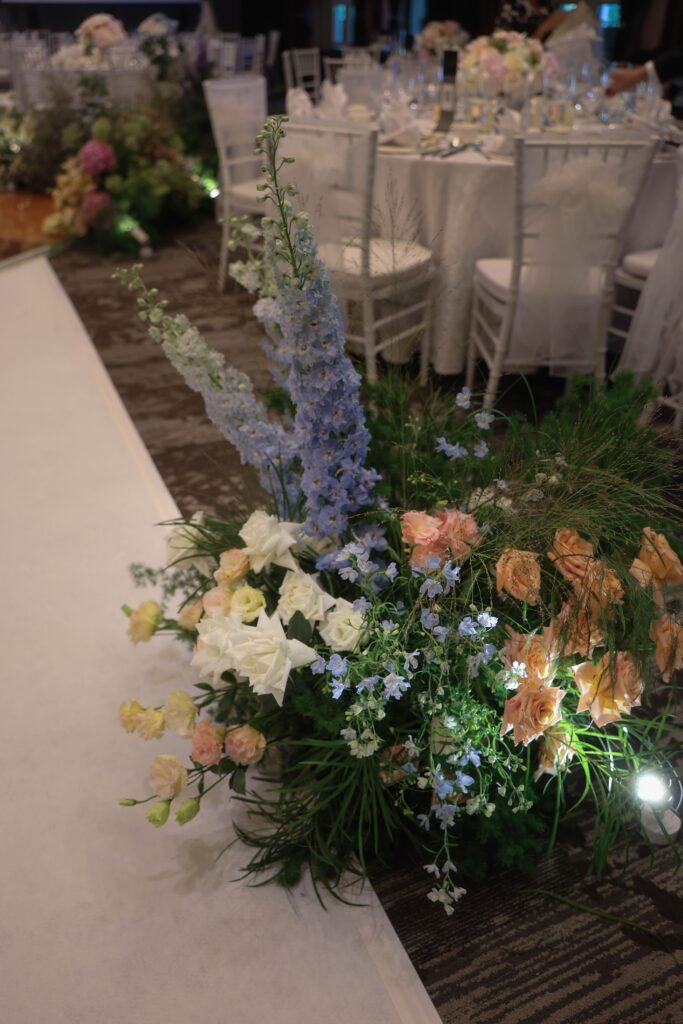 Aisle floral arrangements transitioning into dance floor styling with blue and pink blooms including hydrangeas, roses, and textured greenery in a cohesive romantic layout at The Outpost Hotel