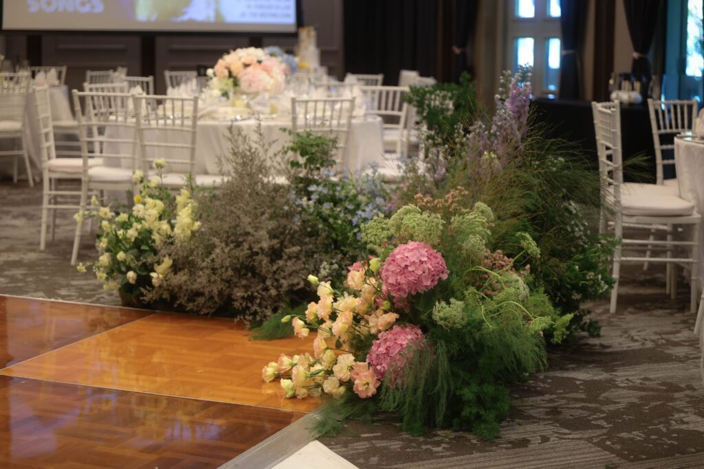 Asymmetrical corner floral arrangement with blue delphiniums, pink roses, hydrangeas, and layered greenery creating a soft garden composition around the dance floor at The Outpost Hotel