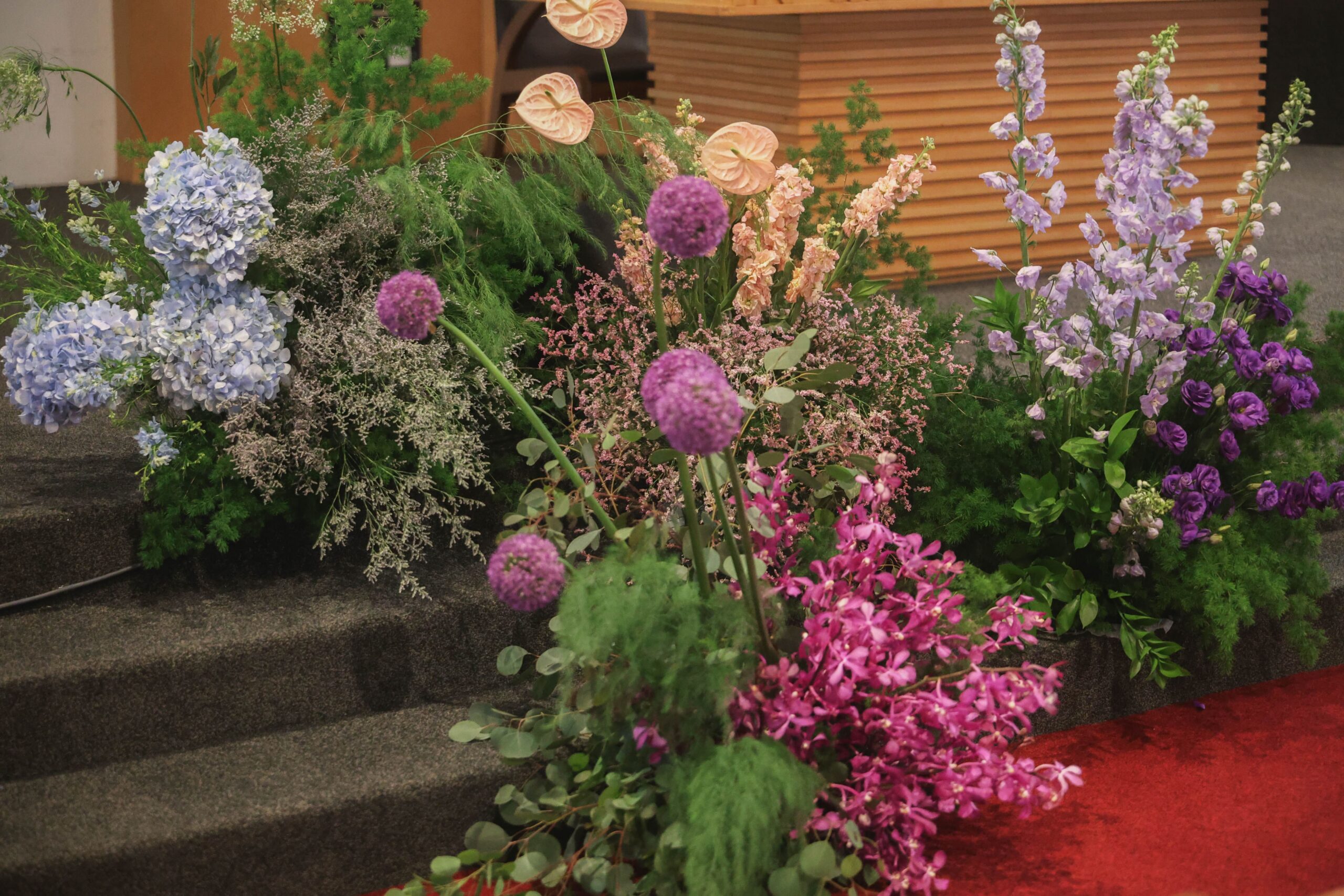 Colourful ceremony floral arrangement featuring lavender Delphinium, blue Hydrangea, purple Giant Allium, pink Mokara orchids, Anthuriums, Lisianthus, and layered foliage inspired by botanical artwork in Singapore.