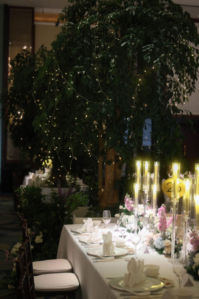Long guest dining table styled with towering illuminated trees, soft candlelight, lush greenery, and romantic floral arrangements in refined ballroom styling at Shangri-La Singapore