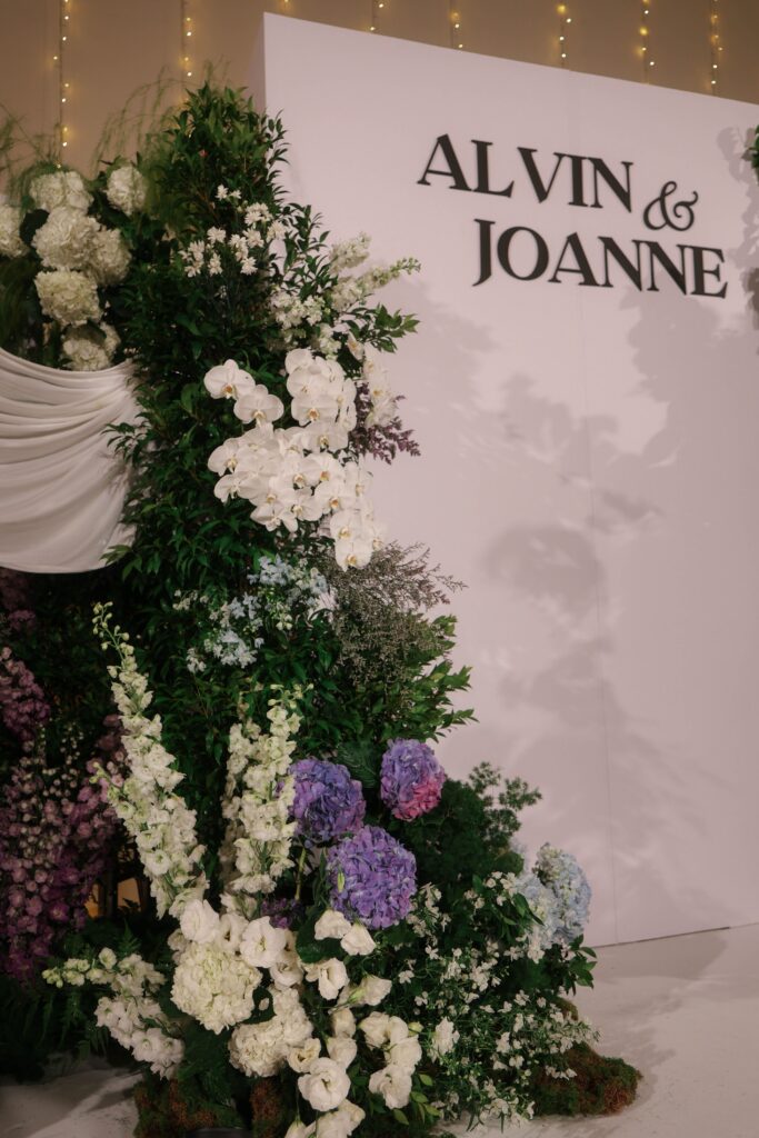 Wedding stage backdrop styled with gathered white drapery, pastel hydrangeas, white florals, soft purple blooms, and organic greenery in elegant ballroom design at Shangri-La Singapore