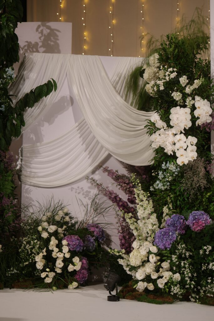Close-up detail of wedding stage drapery styled with layered white fabric, pastel blue and purple floral arrangements, and lush organic greenery at Shangri-La Singapore