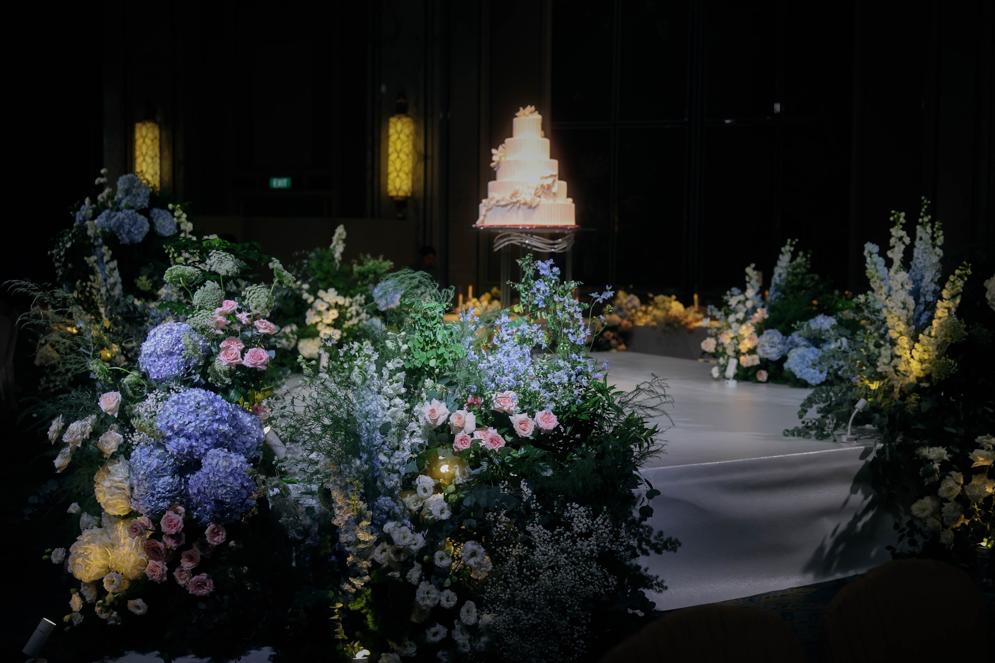 tiered cake on a central stage ringed by a garden of blue hydrangea, blush roses and delphinium