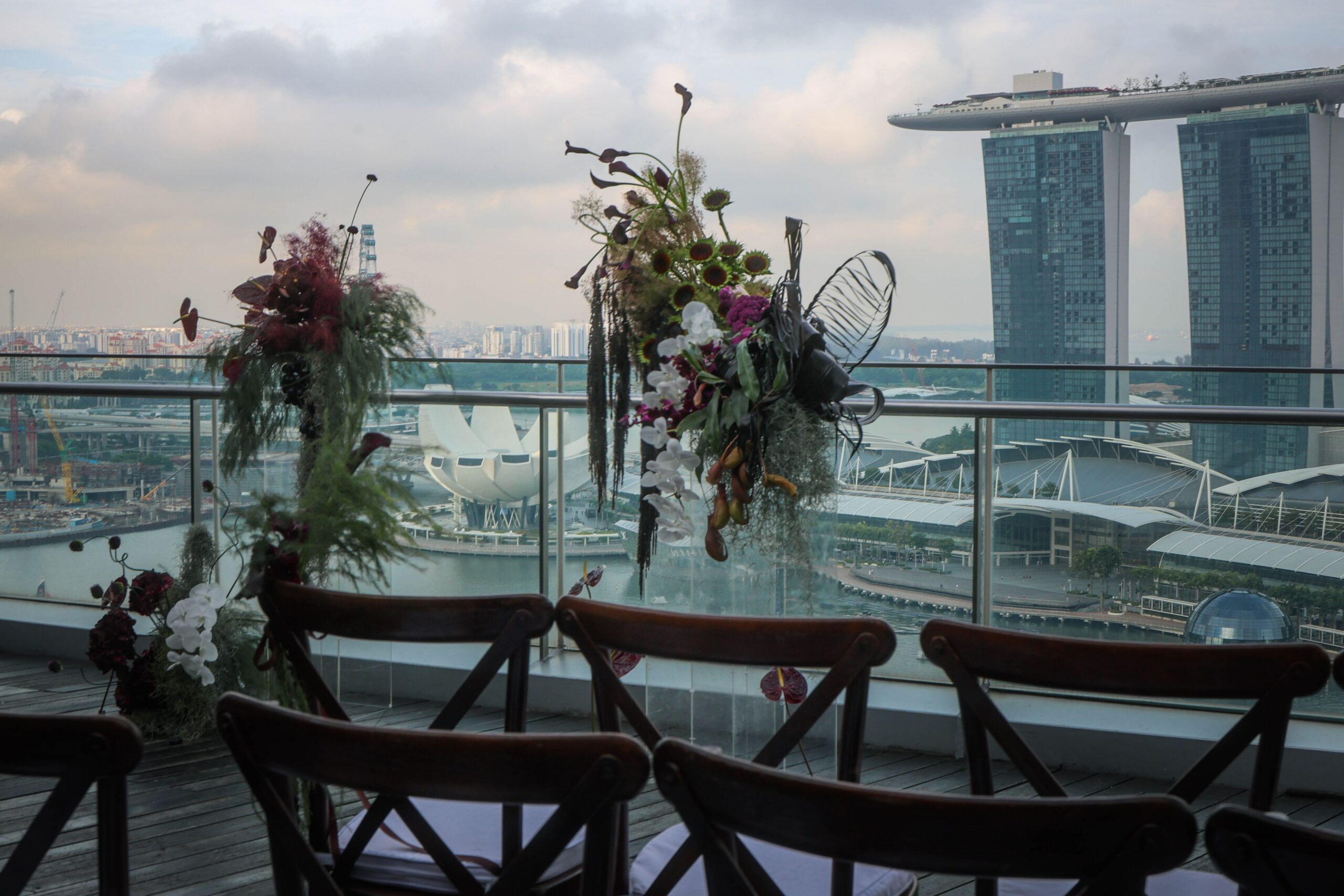 Dark and moody ceremony backdrop and reception floral styling overlooking Marina Bay with sunflowers, orchids, calla lilies, burgundy amaranthus, and sculptural foliage at VUE Singapore.