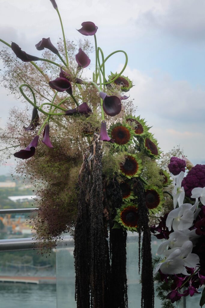 floral installation featuring burgundy calla lilies, sunflowers, white orchids, cascading amaranthus, and textured foliage against the Marina Bay skyline at VUE Singapore.Contemporary floral arrangement featuring orchids, burgundy anthuriums, hydrangeas, and airy botanical textures at VUE Singapore.