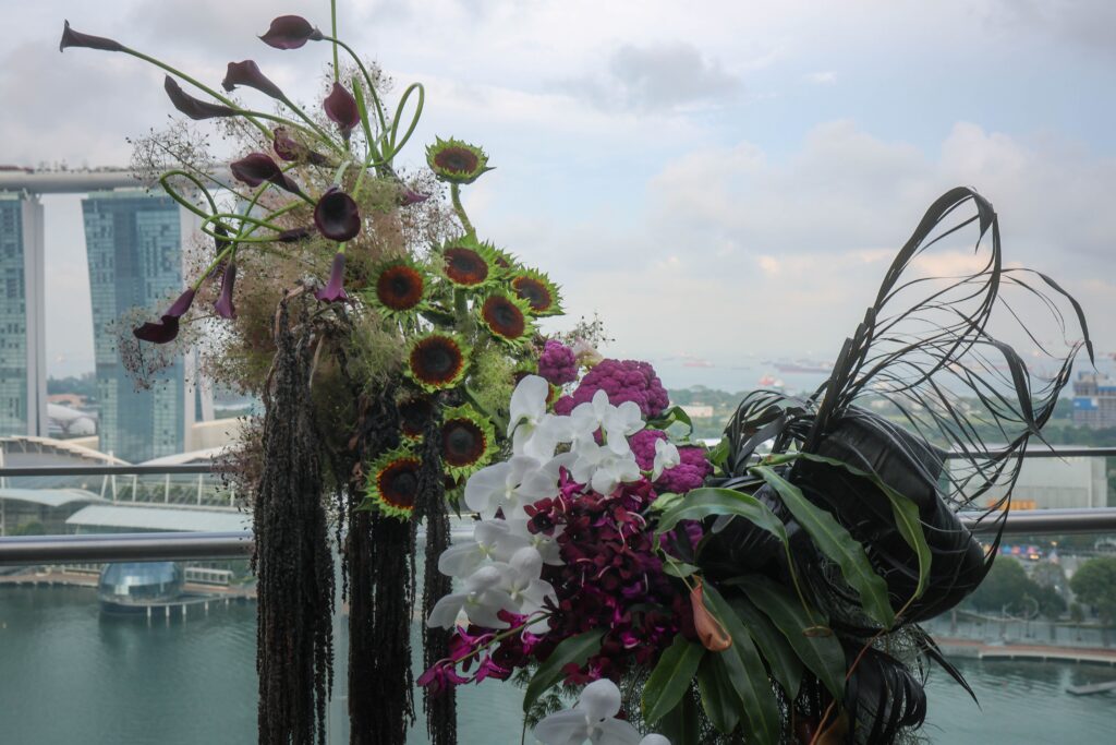 Side view of the ceremony backdrop featuring sunflowers, orchids, calla lilies, cascading amaranthus, and sculptural foliage overlooking Marina Bay at VUE Singapore.