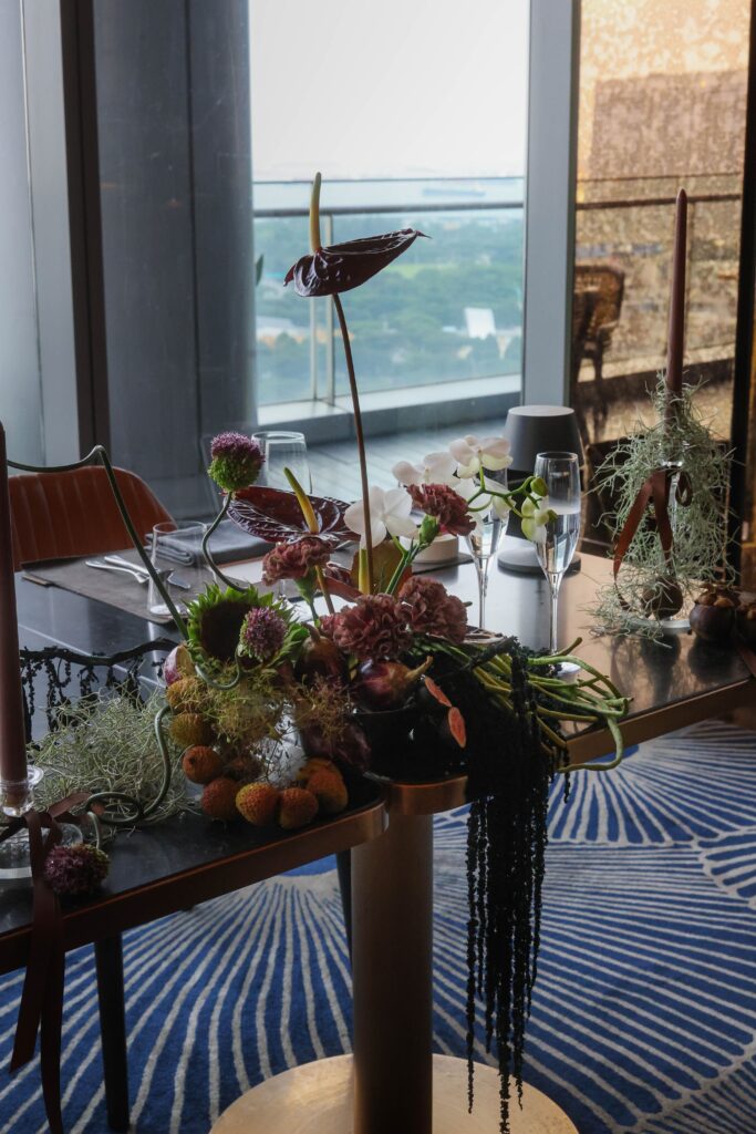 Reception floral arrangement with cascading foliage, athriums, sunfllowers, amaranthus, long beans and rich botanical textures overlooking Marina Bay at VUE Singapore.