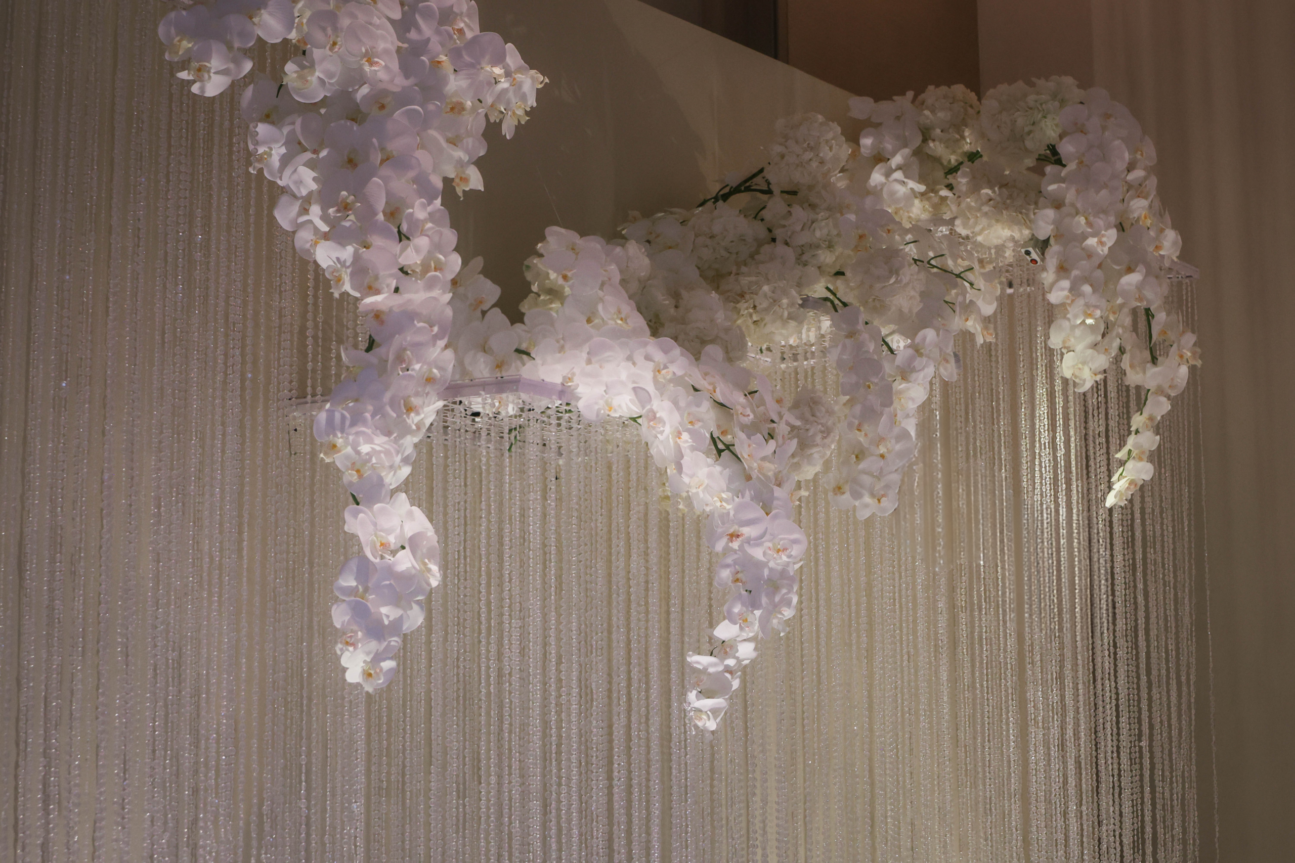 White Phalaenopsis orchids and hydrangeas cascade organically from a hanging crystal wedding backdrop, balancing soft floral forms with structured vertical crystal strands at The Singapore EDITION, Singapore.